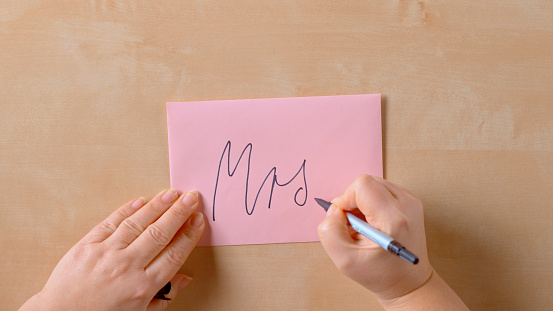 Close-up of woman's hands writing inscription Mrs. on back of pink envelope.