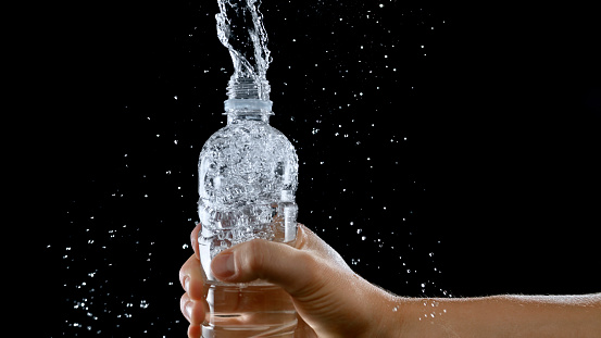Close-up of man's hand holding water bottle splashing water against black background.