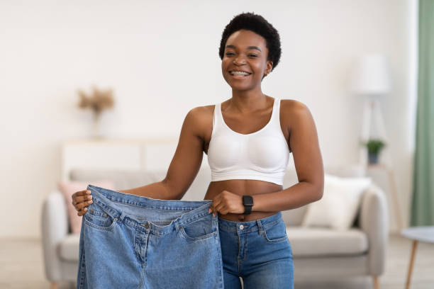 Skinny Black Female Showing Old Large Jeans After Weight-Loss Indoor Slimming Motivation Concept. Skinny Black Female Showing Old Large Jeans After Successful Diet And Weight Loss Posing Smiling To Camera At Home. Weight-Loss Before And After Comparison before and after weight loss stock pictures, royalty-free photos & images
