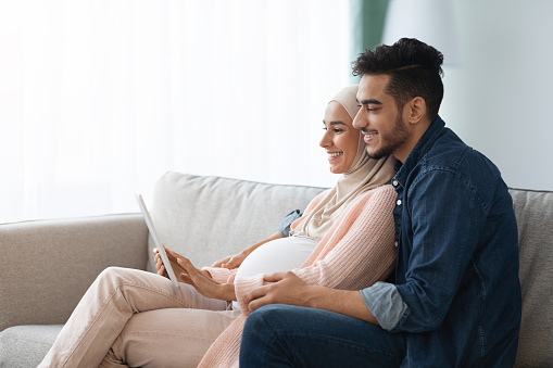 Pregnant Muslim Woman And Her Husband Relaxing With Digital Tablet At Home, Happy Islamic Spouses Shopping Online In Internet Or Browsing Social Networks, Enjoying Spending Time Together, Free Space