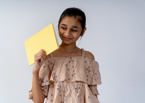 Happy Asian, an Indian teenager girl student holding a book in hand and standing against isolated white background.