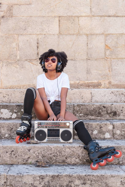 Beautiful black woman with afro hair, roller skates and sunglasses listens to music with headphones from an old radio cassette player while sitting on a stone stairs outdoors on a hot summer day stock photo