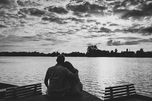Rear view of a romantic man and woman sitting on the pier or bridge enjoying stunning sunset. Concept of lovely family. Young couple hugging and looking away. Black and white photo.