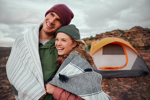 Beautiful woman laughing while hugging man wrapped in a blanket during camping looking at view