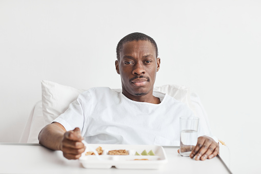 Front view portrait of African-American man eating food in hospital while lying on white bed and looking at camera, copy space