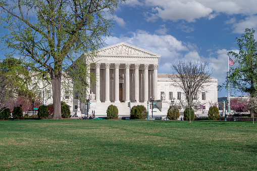 U.S. Supreme Court Building, Washington DC, USA. Green lawn, trees in bloom and bushes are in foreground. Blue sky with Puffy Clouds is in background. \n.