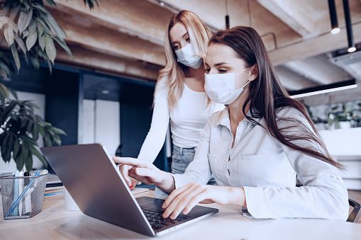 Two female colleagues working in office together wearing medical masks, coronavirus prevention