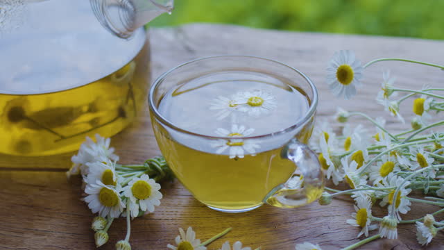 Chamomile tea in a glass teapot and cup, the camera moves around. The action takes place on a summer rural terrace with playing sunbeams. Nearby are the flowers of a pharmaceutical chamomile.