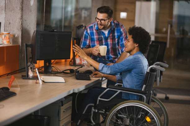 behinderter computerprogrammierer sitzt im rollstuhl am arbeitsplatz und arbeitet mit einem kollegen zusammen - working physical impairment people occupation stock-fotos und bilder