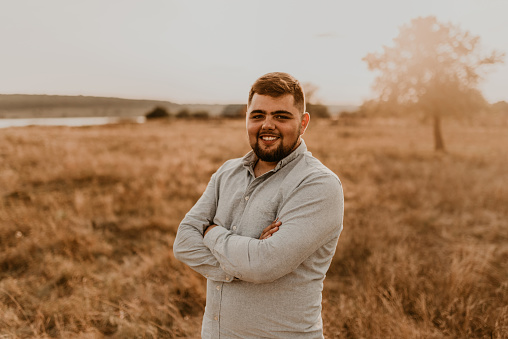 young caucasian black-haired overweight man with beard smiling cheerfully in summer cotton clothes and shorts outdoor in sunny weather. men fashion collection. open spaces meadow flooded sunlight tree