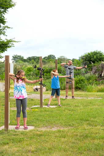 Two young girls and their dad practicing at an outdoor archery range on an overcast summer day. Focus is on the girl nearest the camera, but her younger sister and dad can be seen beyond her, all drawing their bows to shoot at their targets.