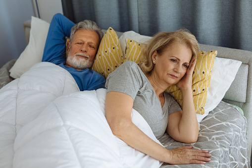 Irritated senior woman blocking ears while man snoring in bed