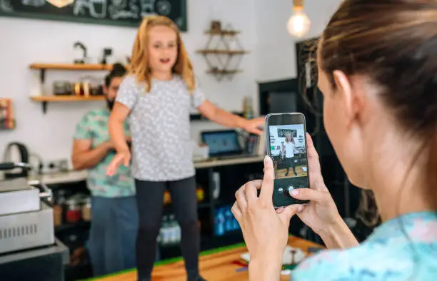 Mother recording her daughter dancing with the mobile while working in a coffee shop. Reconciliation family life work concept. Selective focus on mobile in foreground