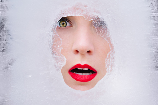 Beautiful young model face in ice. Cold frozen winter beauty with red lips in ice cube. Fashion art woman portrait. High quality photo