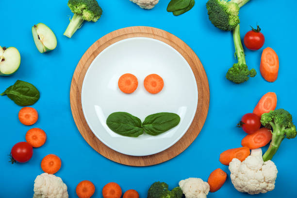 istock Plate and smiley food on blue background surrounded by fruits and vegetables. Complementary feeding for children, food allergy, diet, healthy eating concept. Spinach, broccoli, cauliflower, carrot, apple. 1312685481