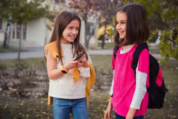 Photo of Silly young girls holding smart phone and looking at her happy friend