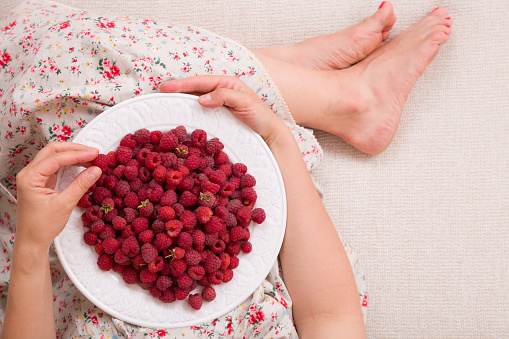 Top view on woman sitting cozy on the couch and enjoying delicious summer berries. Girl holding white plate with organic raspberries on her knees