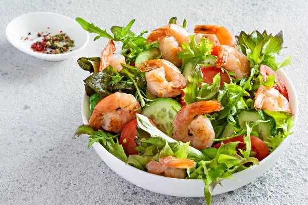 Photo of Fresh crispy salad with vegetables and seafood (shrimps) in a white plate on a light background.