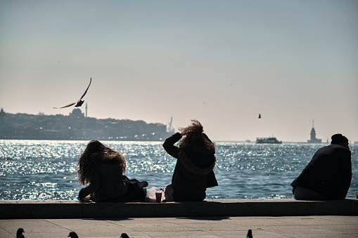 Turkey istanbul 04.03.2021. Girls sitting shore of the bosporus. curly-haired women and her hairs waves from winds with seagulls, ship and ferry background during morning and sun reflection on sea.