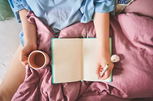 High angle shot of an unrecognizable woman sitting alone at home and holding a cup of coffee while journaling