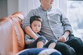 Asian chinese father spending time with his son in living room sitting on sofa watching tv