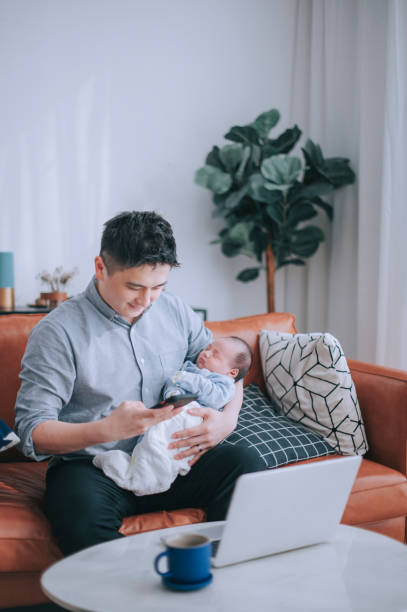 istock asian chinese man working from home reading text message from smart phone while carrying his baby boy sitting on sofa with laptop in living room 1312722895