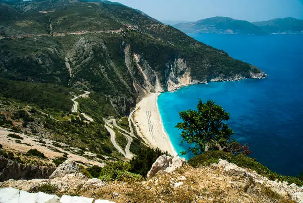 Myrtos Beach from the cliff top illustrates lazy days under cooling breezes by warm deep turquoise Mediterranean sea, surrounded by green hills covered with vegetation. Kephalonia is one of the greenest islands in Greece.