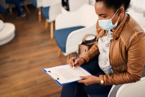 African American woman with face mask filling out medical papers at dental waiting room. Black woman writing her data in medical document before dental examination. She is wearing protective face mask due to COVID-19 pandemic. medical insurance stock pictures, royalty-free photos & images