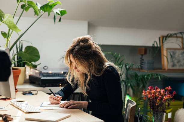 Young Caucasian Female Professional Working from Home- Sitting in her Office Area and Writing Notes (Horizontal) Beautiful blond woman making a to do list while sitting behind a desk in her living room writing activity stock pictures, royalty-free photos & images