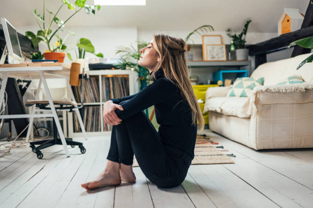 New Normal Concept: Young Beautiful Caucasian Woman Spending her Free Time Alone in her Studio Apartment Horizontal side view shot of a beautiful blond woman dressed in black, sitting on a floor in the middle of a bright living room, practicing breathing with her eyes closed (copy space) inhaling stock pictures, royalty-free photos & images