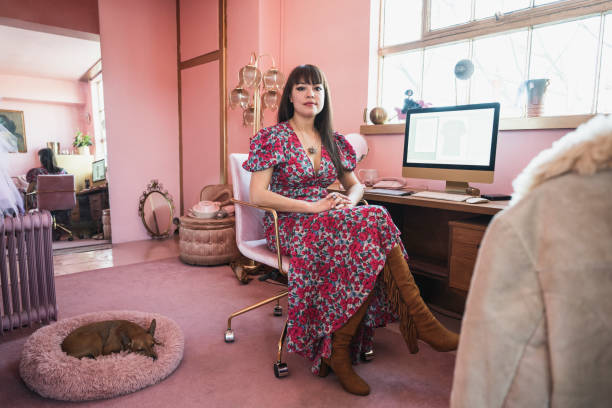 Working portrait of early 40s design professional in atelier Full length view of mixed race woman wearing floral pattern dress and fringed boots sitting in office chair and smiling at camera with sleeping dog by her side. one mature woman only stock pictures, royalty-free photos & images