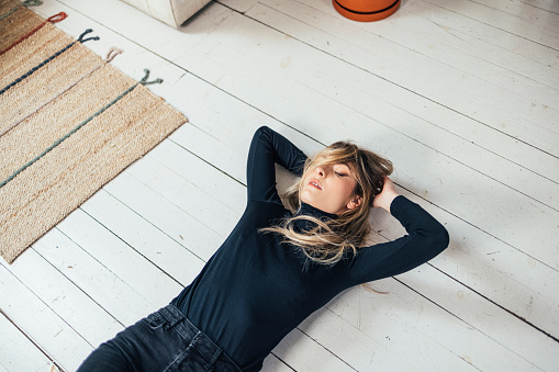 Wide angle shot of a tired young woman lying on a white wooden floor with messy, eyes closed and arms under her head (copy space)