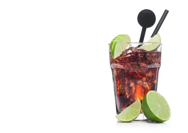 Cuba Libre Cocktail in glass with ice cubes and slices of lime with black straw and stirrer on white background with raw limes.Space for text