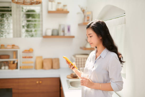 istock Beautiful happy girl drinking water while using mobile phone at home kitchen 1312777475