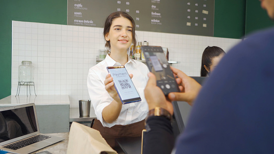 Young Middle East woman barista people in apron collecting an order payment by scanning QR code on phone screen, talking to customer at the counter, coffee shop cafe cashier. People lifestyle. Service