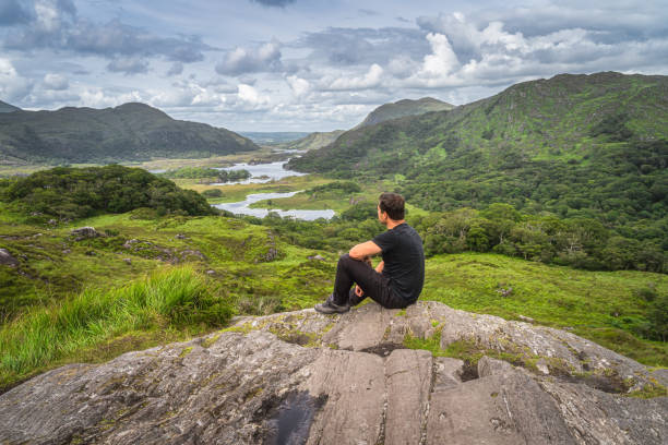 Middle age man sitting on a rock and admiring beautiful Ladies View Middle age man sitting on a rock and admiring beautiful Ladies View, one of iconic Irish viewpoints, Lakes of Killarney, Rink of Kerry, Ireland killarney lake stock pictures, royalty-free photos & images