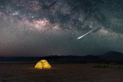 Yellow tent under Milky way galaxy with meteor in desert in the countryside of northern Thailand, Stars above the night mountain forest and a glowing camping tent.