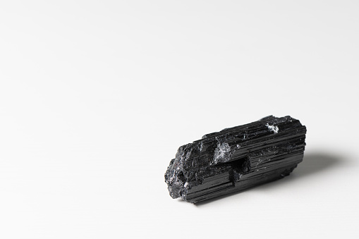 This is a photograph of a raw black tourmaline log on a white background. Tourmaline is a semi-precious gemstone that is used energetically for balancing and grounding purposes.