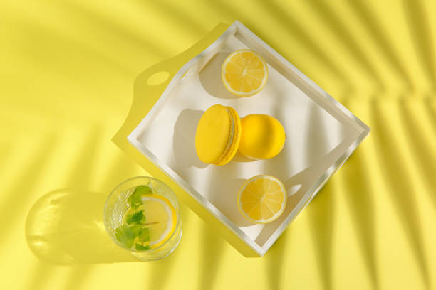 Yellow macaroon with lemons on white tray and glass of lemonade on background with palm tree shadow. Summer refreshment concept. Top view.