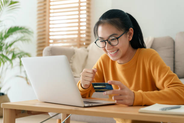 belle jeune femme achètent en ligne avec une carte de crédit tout en s’asseyant dans le matin de salon. les femmes utilisent un ordinateur portable et font des transactions en ligne à la maison. - achats en ligne photos et images de collection