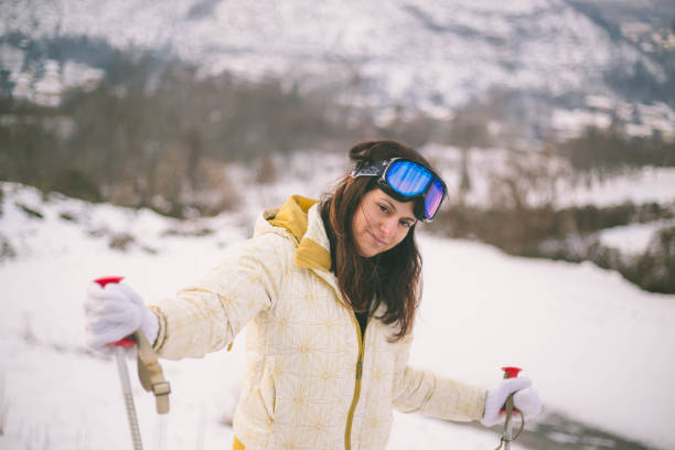 Close-up view beautiful young woman in winter clothes stands sideways and holds sticks in hand on a background the snow mountain
