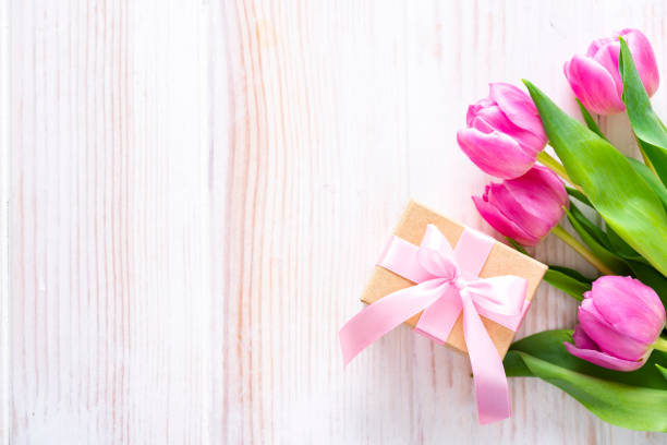 Top view of a brown gift box and pink tulips arranged at the right of a white table frame leaving useful copy space for text and/or logo at the left. Predominant colors are white and pink. High resolution 42Mp studio digital capture taken with Sony A7rII and Sony FE 90mm f2.8 macro G OSS lens