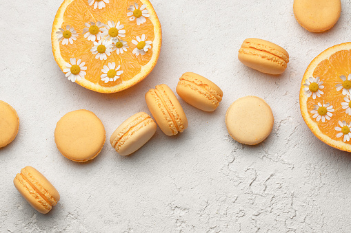 Orange French Macaroons with orange fruit filling on a creamy white clay background, decorated with small chamomiles and fresh sliced oranges