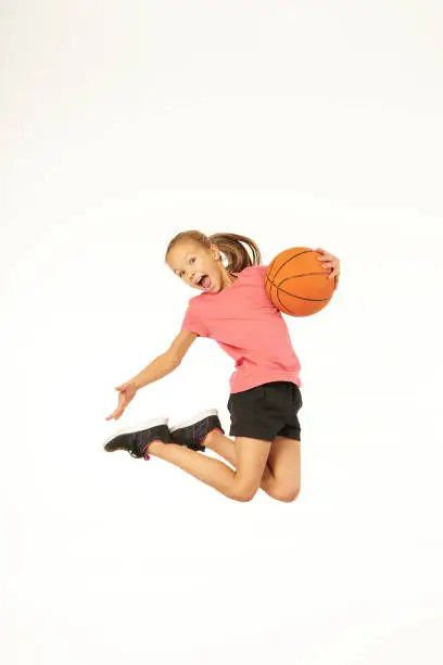 Cute female child basketball player holding game ball and screaming while jumping in the air. Isolated on white background