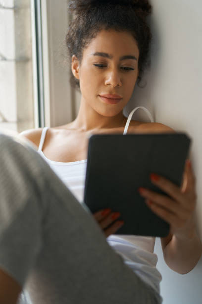 istock Relaxed young woman reading a book using tablet pc while sitting by the window at home 1312862372