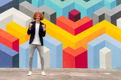 Full body young ginger man with curly hair gesturing horns while standing near wall with colorful geometric ornament on street
