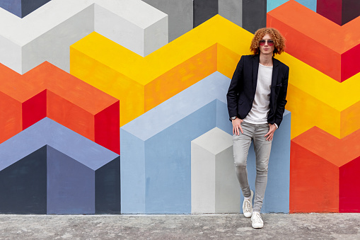 Full body of contemporary young redhead male in sunglasses and smart casual style clothing leaning against wall with colorful geometric ornament and looking at camera