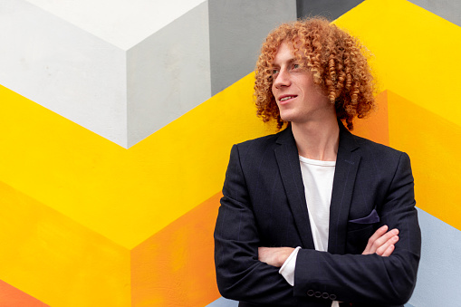 Positive young curly haired ginger male in stylish casual outfit with arms crossed smiling and looking away thoughtfully while standing against colorful geometric background