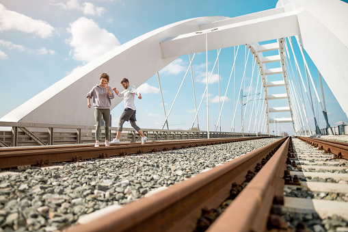 Photo of sporty young couple having fun next to train tracks on the bridge. They are running on railroad and having a race.