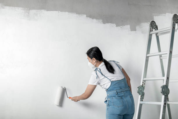 chica pinta una pared blanca con un rodillo. reparación del interior. joven decoradora femenina pintando una pared en la habitación vacía, creadora de conceptos o pintora con casco con rodillo de pintura sobre la habitación vacía. - female house painter home decorator paintbrush fotografías e imágenes de stock